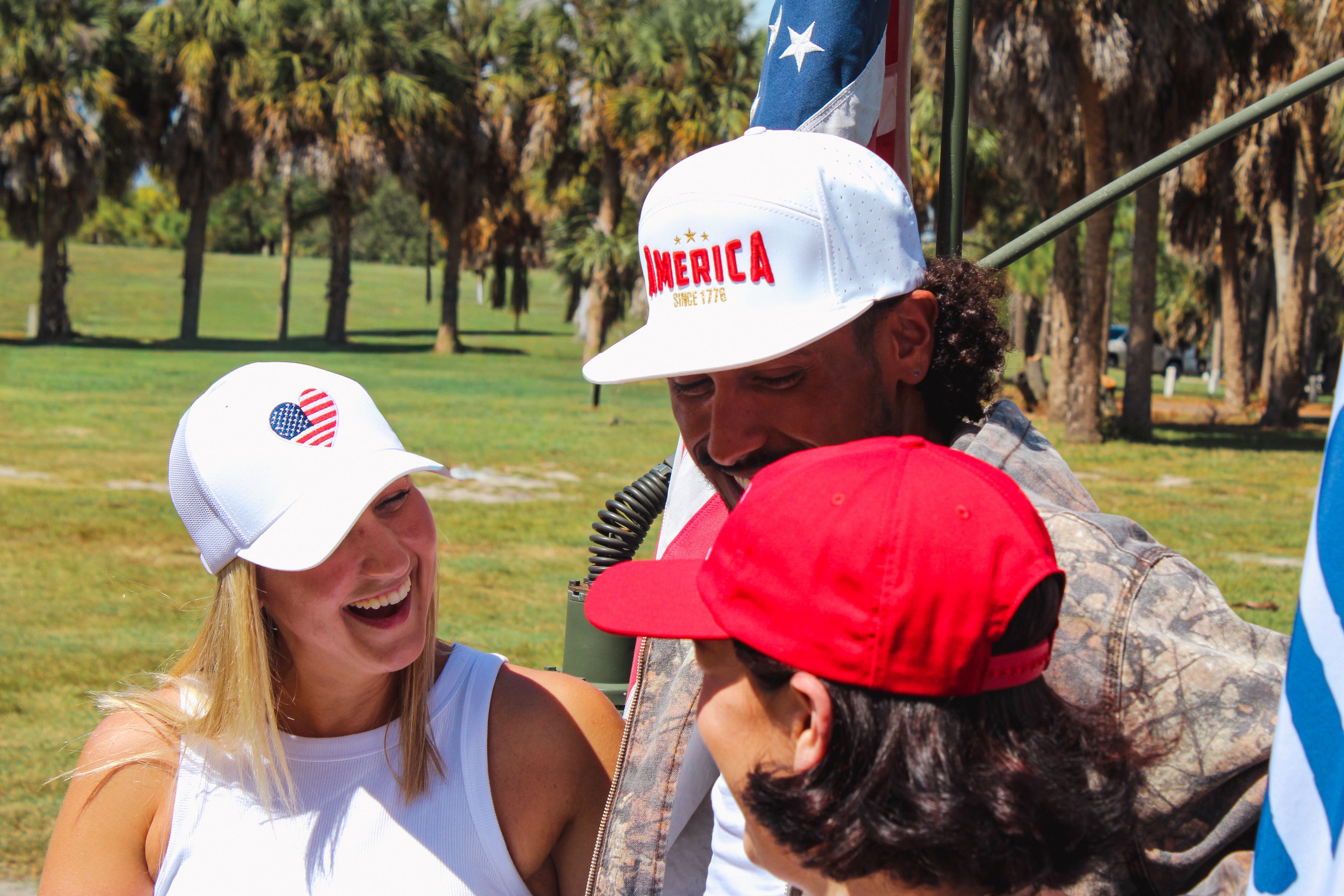 Three people wearing American themed hats with text and flags on a grassy field.